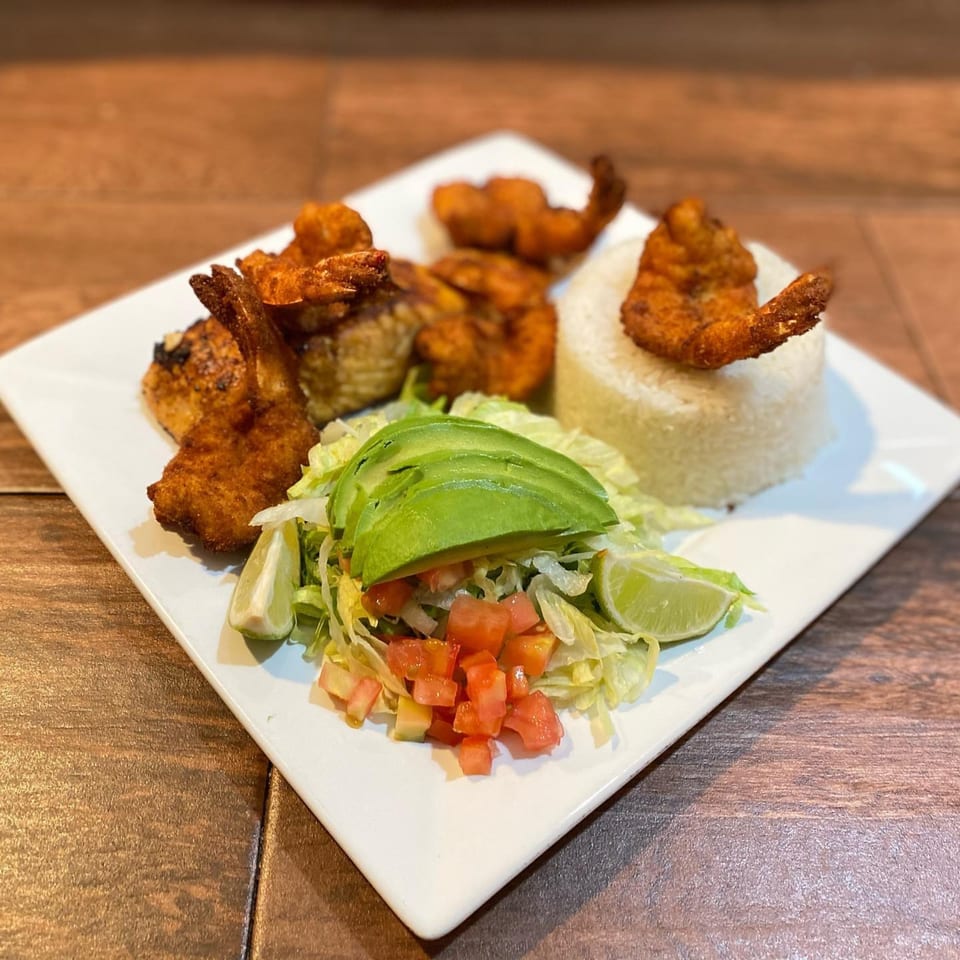 Fried shrimp and chicken with rice, lettuce, avocado, diced tomatoes, and lime on a white plate.