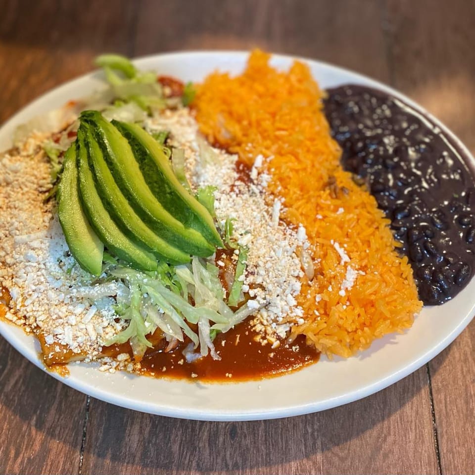 Mexican enchiladas topped with avocado, lettuce, and cheese, served with orange rice and black beans.
