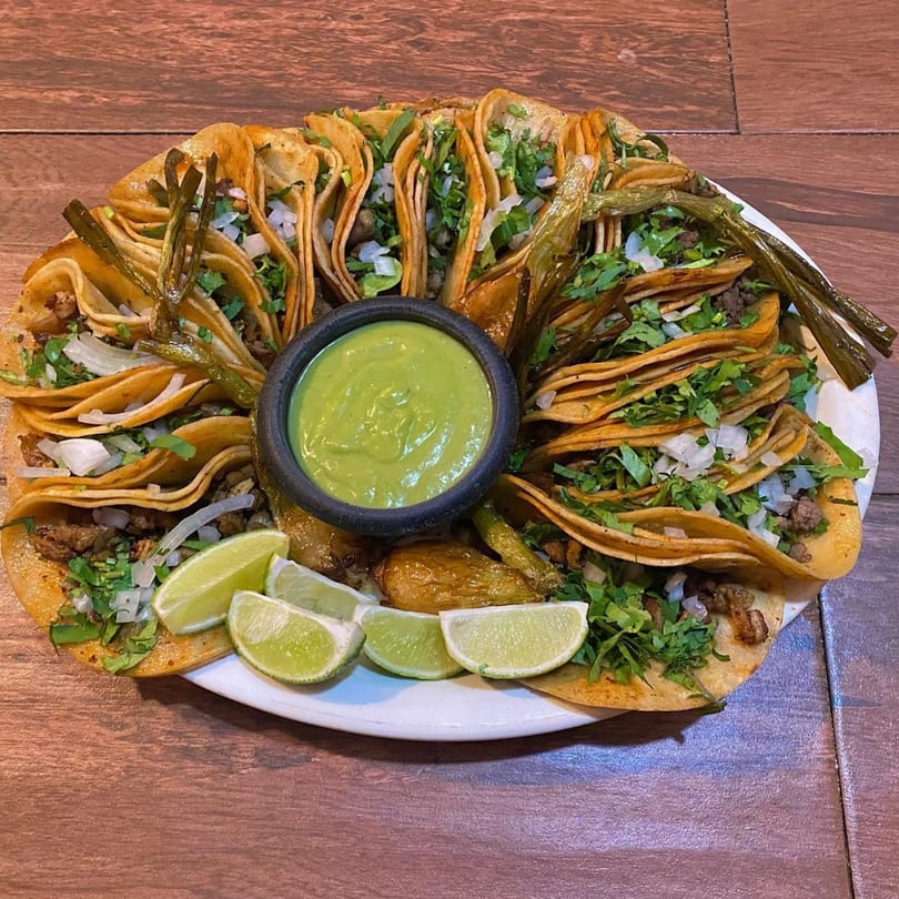 A large platter of El Pastor tacos with green salsa, grilled peppers, and lime wedges.