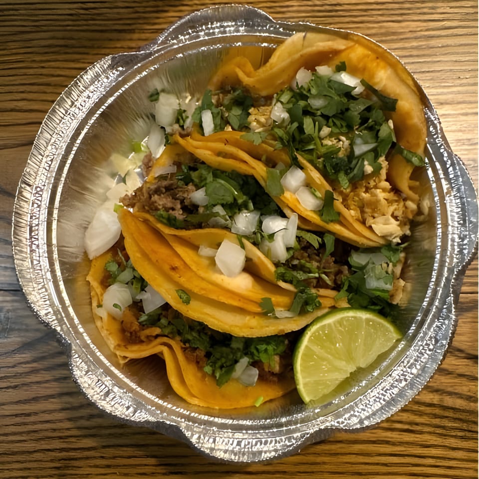 Three El Pastor tacos in a foil container, topped with cilantro, onions, and a lime wedge.