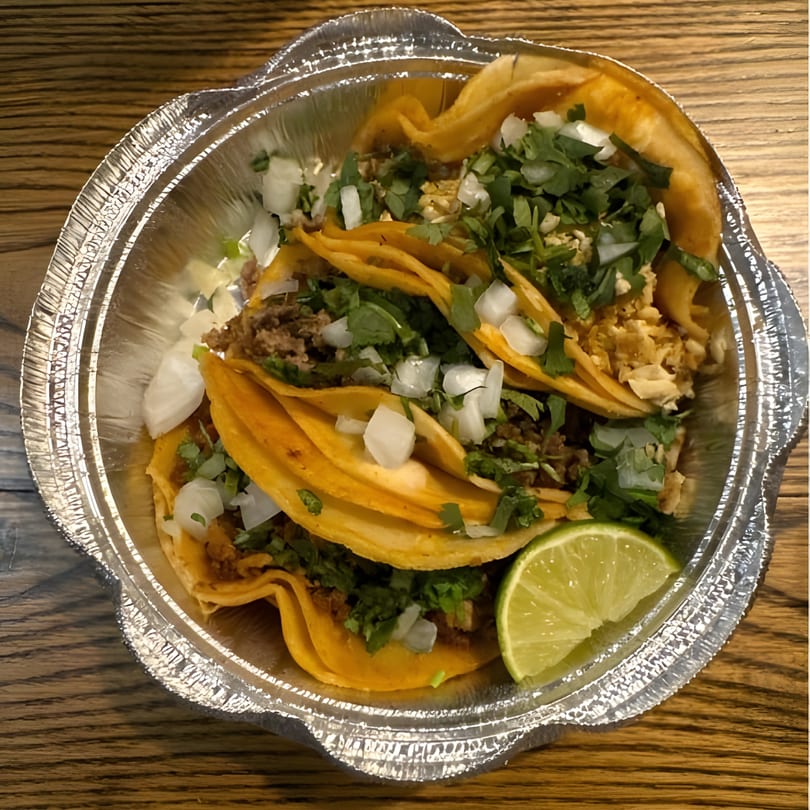Three El Pastor tacos in a foil container, topped with cilantro, onions, and a lime wedge.