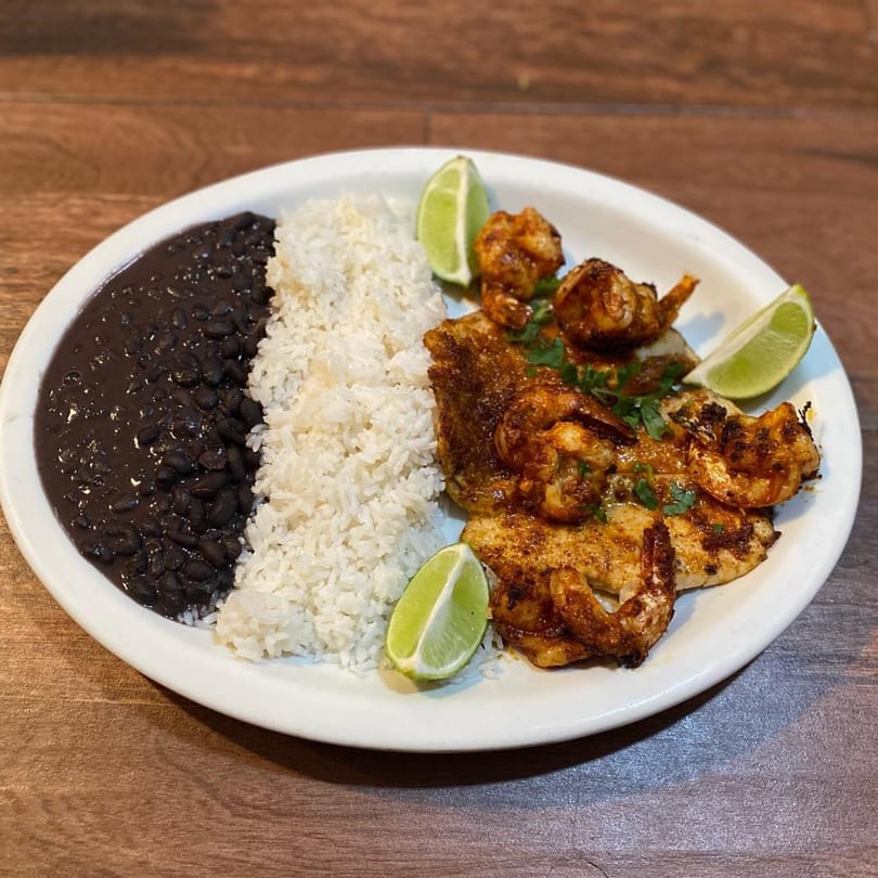El Pastor's grilled chicken and shrimp with rice, black beans, and lime wedges on a white plate.
