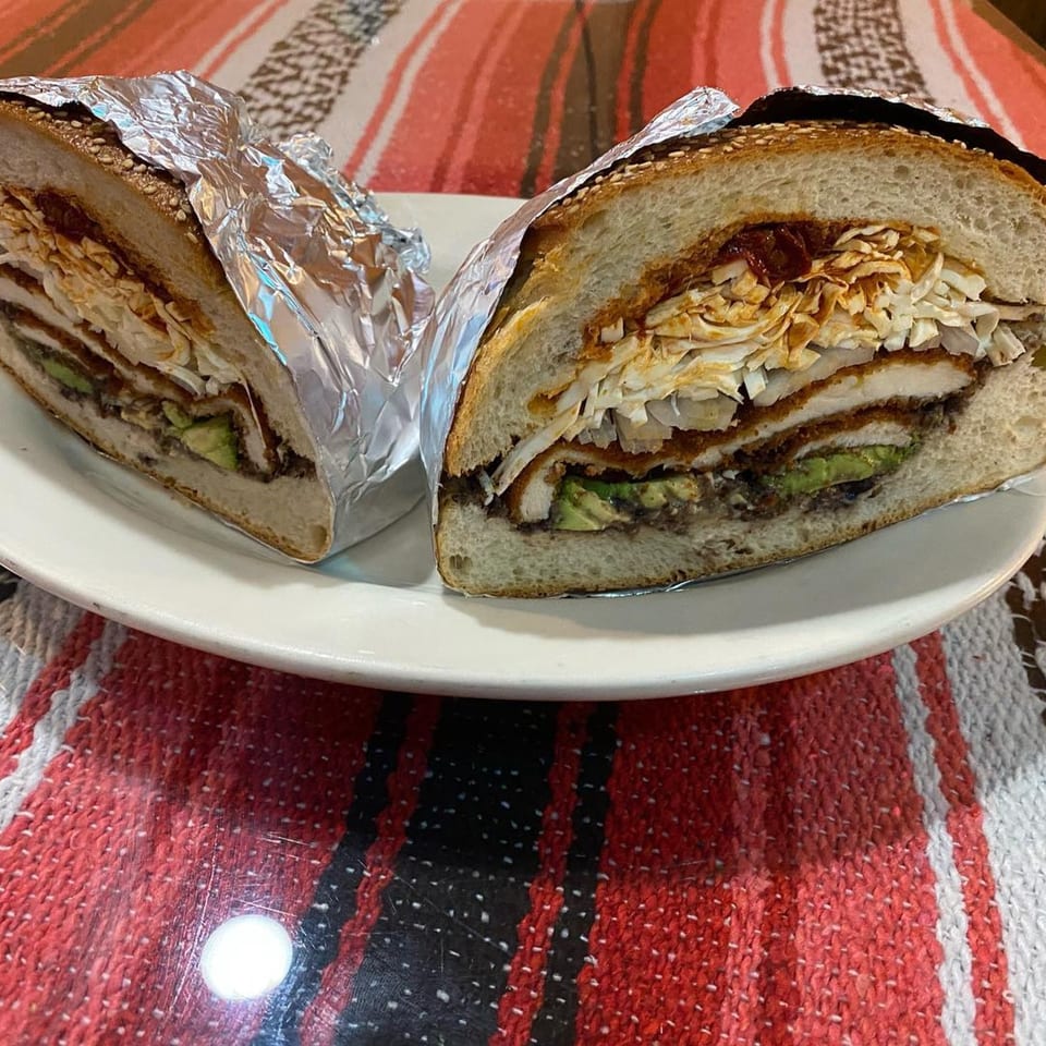 Two halves of a large Mexican torta sandwich, filled with meat, avocado, and cabbage, on a white plate.