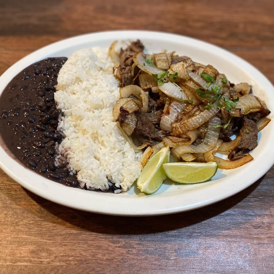 Mexican dish with white rice, black beans, steak and onions, garnished with lime wedges and cilantro.