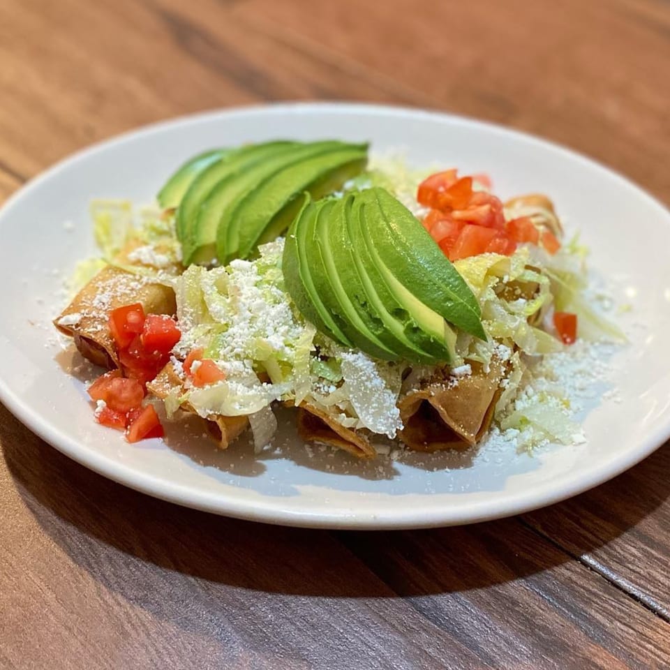 Crispy taquitos topped with shredded lettuce, diced tomatoes, crumbled cheese, and sliced avocado.